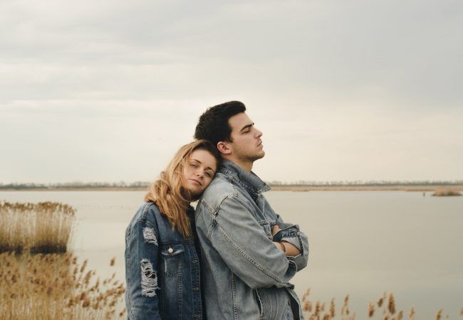 woman in blue denim jacket leaning on man's shoulder near body of water